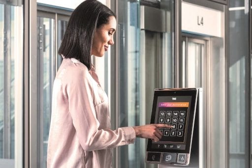 Eine Frau steht seitlich zum Aufzug  im 4. Stockwerk und drückt auf einem Touchpad die Taste 6. 