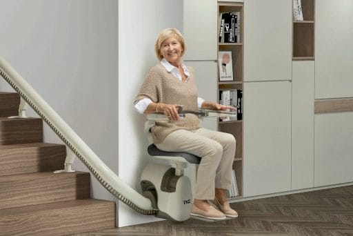 Eine Frau mittleren Alters mit blonden Haaren sitzt auf einem Sitzlift am unteren Ende der Treppe. Ihr Blick ist in die Kamera gerichtet.